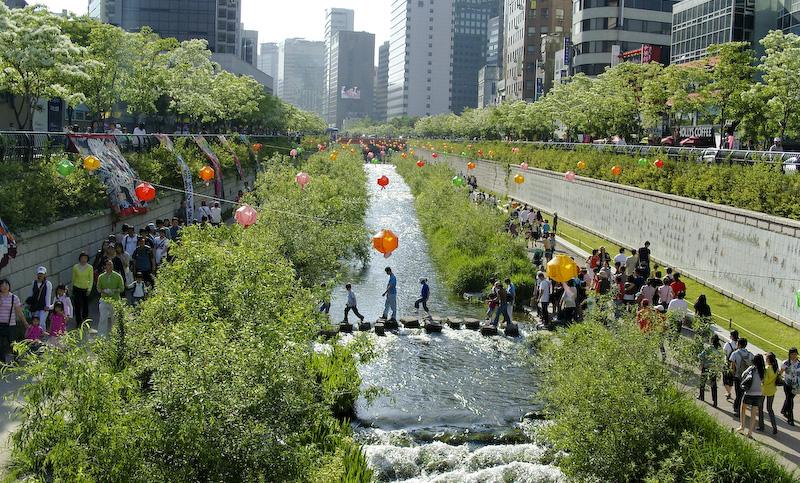 People stroll on two paths following a waterway bordered by greenery. A few folks cross a bridge over the stream.